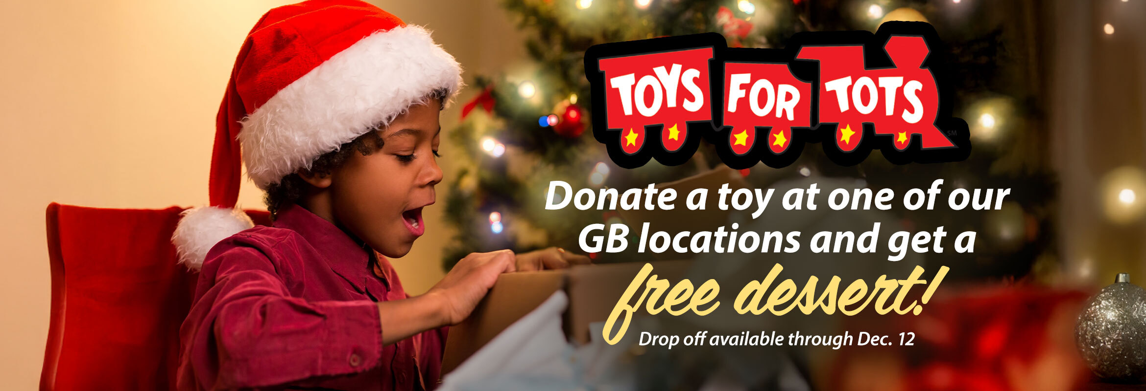 child in Santa hat looking into a box and smiling with wonder, with Christmas tree in background